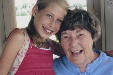 A young blond girl in a pink sleeveless top stands next to a woman with short brown hair and a light blue top. The girl has her arm around the woman's shoulders.